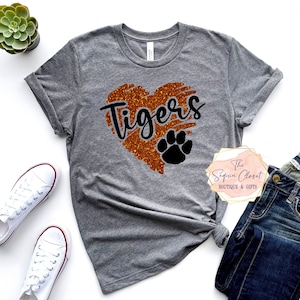 May include: Gray t-shirt with a glittery orange heart that says "Tigers" in black lettering. A black paw print is inside the heart. The t-shirt is on a white background with a pair of white sneakers and blue jeans.