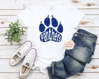 May include: White t-shirt with a blue glitter paw print and the word "Panthers" inside the print. The t-shirt is laying on a white wooden surface with a pair of white sneakers and blue jeans.