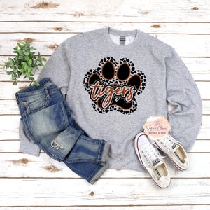 May include: Gray sweatshirt with a brown and black paw print design. The paw print has the word "tigers" written inside.