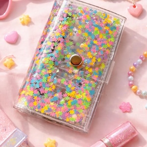 Könnte beinhalten: Transparente Geldbörse mit bunten Stern-Glitzer-Füllung und goldenem Druckknopfverschluss. Das Bild zeigt rosa herzförmige Sonnenbrillen, Lipgloss und Perlenarmbänder. Die Accessoires liegen auf einer rosa Oberfläche.