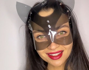 May include: A transparent cat-eye mask with attached ears. The mask is dark gray and secured with silver rivets. The person is wearing a gold chain necklace with a heart pendant and has red lipstick.