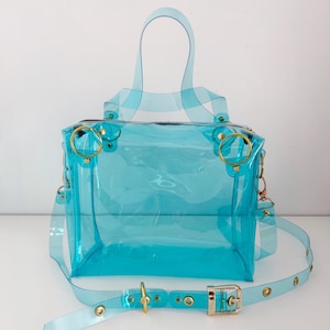 May include: A transparent, turquoise handbag with a top handle and a detachable shoulder strap. The bag features gold-colored hardware, including rings and a buckle. The clear design allows visibility of the contents.