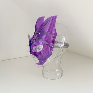 Op de afbeelding: Paarse plastic maskerbalmasker met een geometrisch ontwerp en zilveren studs. Het masker bevindt zich op een mannequinhoofd.