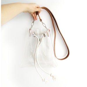 May include: A clear transparent bucket bag featuring a tan leather shoulder strap, silver-toned metal hardware, and a white braided rope drawstring closure with knotted ends. The bag has a structured base and a simple, open-top design.