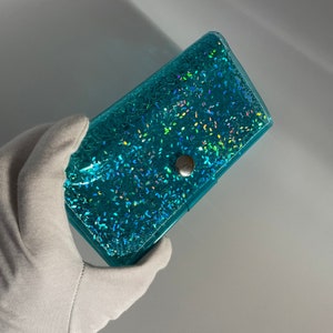 Elegante billetera de vinilo azul con confeti plateado y brillo holográfico. Tiene capacidad para 12 tarjetas e incluye un monedero. Es duradera y fácil de limpiar.