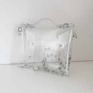 May include: A clear, transparent handbag with a top handle and a detachable shoulder strap. The bag features silver-colored hardware, including rivets and clasps. The design is simple and modern, offering a unique, see-through aesthetic.