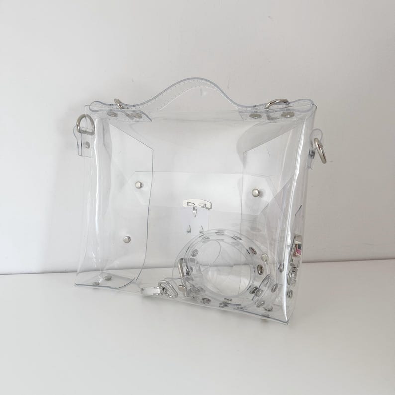 May include: A clear, transparent handbag with a top handle and a detachable shoulder strap. The bag features silver-colored hardware, including rivets and clasps. The design is simple and modern, offering a unique, see-through aesthetic.