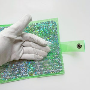 May include: A transparent green wallet with a snap closure. The interior is filled with iridescent glitter. The wallet is being held by a gloved hand.