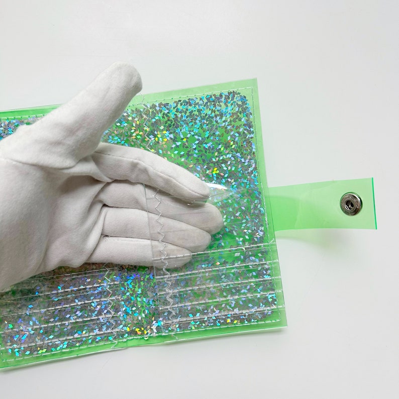May include: A transparent green wallet with a snap closure. The interior is filled with iridescent glitter. The wallet is being held by a gloved hand.