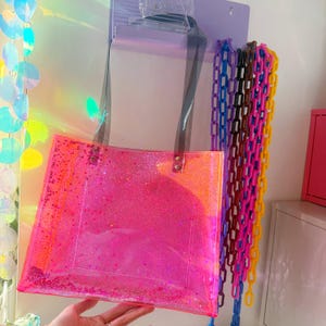 Puede incluir: Un bolso tote transparente con un dise&ntilde;o brillante rosa vibrante. El bolso tiene correas negras y est&aacute; colgado en una pared. Cadenas de pl&aacute;stico de colores en azul, morado, rosa y amarillo cuelgan junto al bolso.