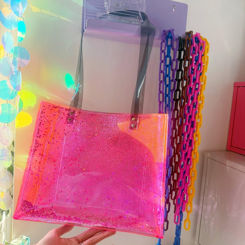 Puede incluir: Un bolso tote transparente con un dise&ntilde;o brillante rosa vibrante. El bolso tiene correas negras y est&aacute; colgado en una pared. Cadenas de pl&aacute;stico de colores en azul, morado, rosa y amarillo cuelgan junto al bolso.