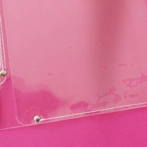 May include: A clear plastic document protector with a pencil inside. The pencil is dark blue with a gold band and eraser. The protector is attached with silver rivets and is on a pink background.