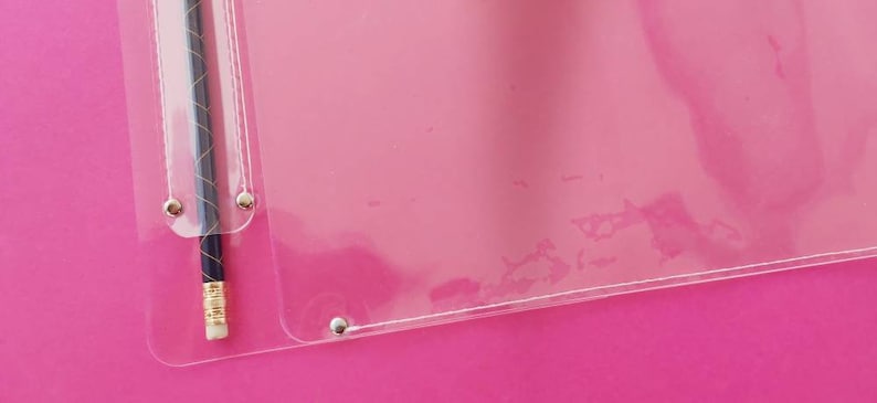 May include: A clear plastic document protector with a pencil inside. The pencil is dark blue with a gold band and eraser. The protector is attached with silver rivets and is on a pink background.
