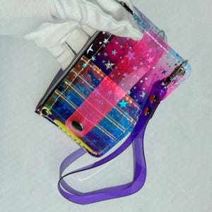 May include: A rectangular, transparent vinyl pouch featuring a vibrant gradient of blue, pink, and yellow hues with a scattered holographic star pattern. The pouch includes a pink snap-button closure and a detachable, translucent purple wrist strap.