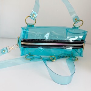 May include: A transparent, turquoise handbag with a rectangular shape. It features a black zipper, gold-colored rings, and a detachable shoulder strap. The bag has a top handle and a crossbody strap, offering versatile carrying options.