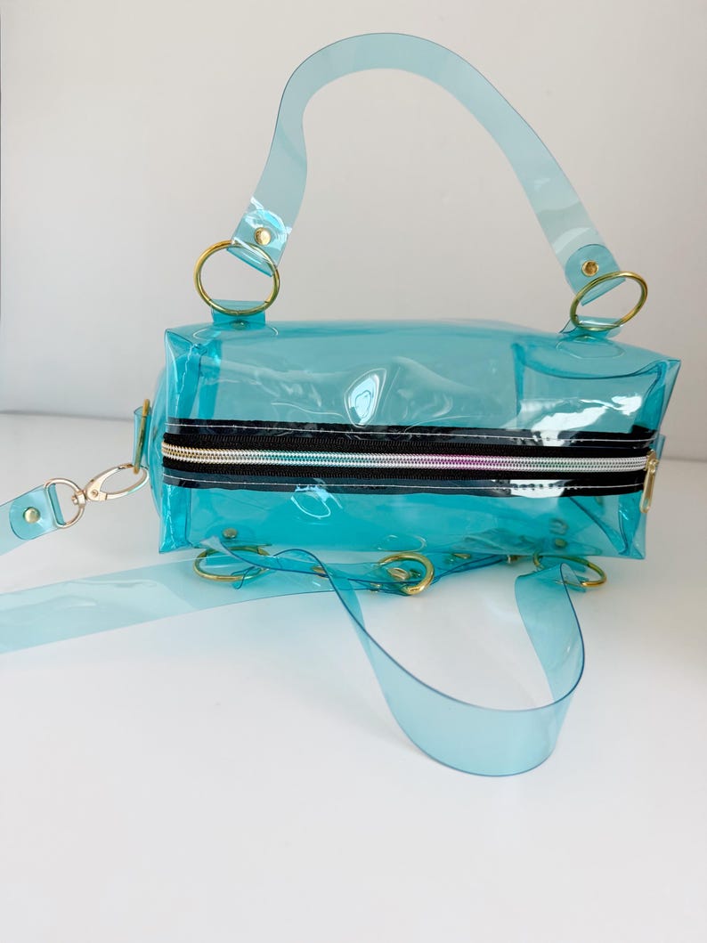 May include: A transparent, turquoise handbag with a rectangular shape. It features a black zipper, gold-colored rings, and a detachable shoulder strap. The bag has a top handle and a crossbody strap, offering versatile carrying options.