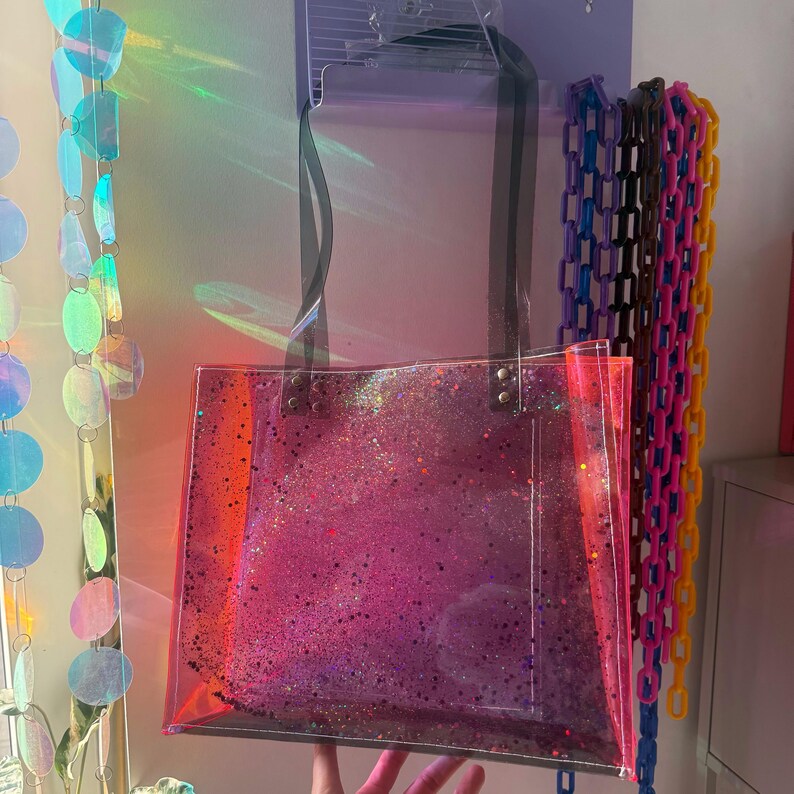Puede incluir: Un bolso tote transparente con un dise&ntilde;o de purpurina rosa. El bolso tiene asas negras y es de forma rectangular. La purpurina se concentra en el centro. El bolso cuelga frente a una pared con decoraciones coloridas.