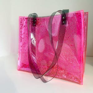Puede incluir: Un bolso tote rosa transparente lleno de purpurina rosa y confeti en forma de estrella. El bolso tiene asas de color gris oscuro y est&aacute; hecho de un material transparente y flexible. El bolso est&aacute; sobre una superficie blanca.