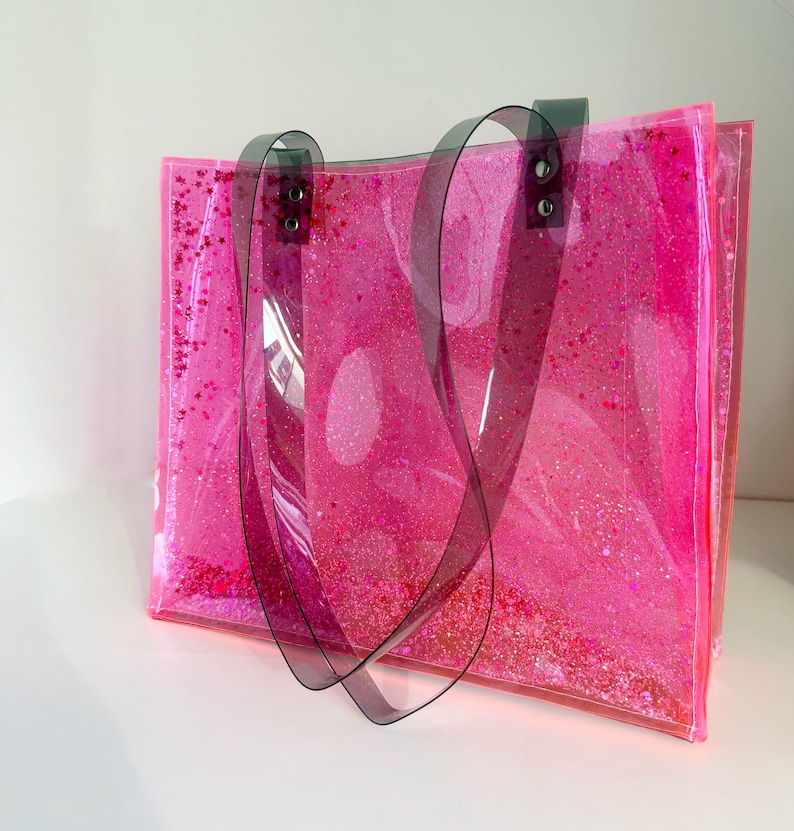 Puede incluir: Un bolso tote rosa transparente lleno de purpurina rosa y confeti en forma de estrella. El bolso tiene asas de color gris oscuro y est&aacute; hecho de un material transparente y flexible. El bolso est&aacute; sobre una superficie blanca.