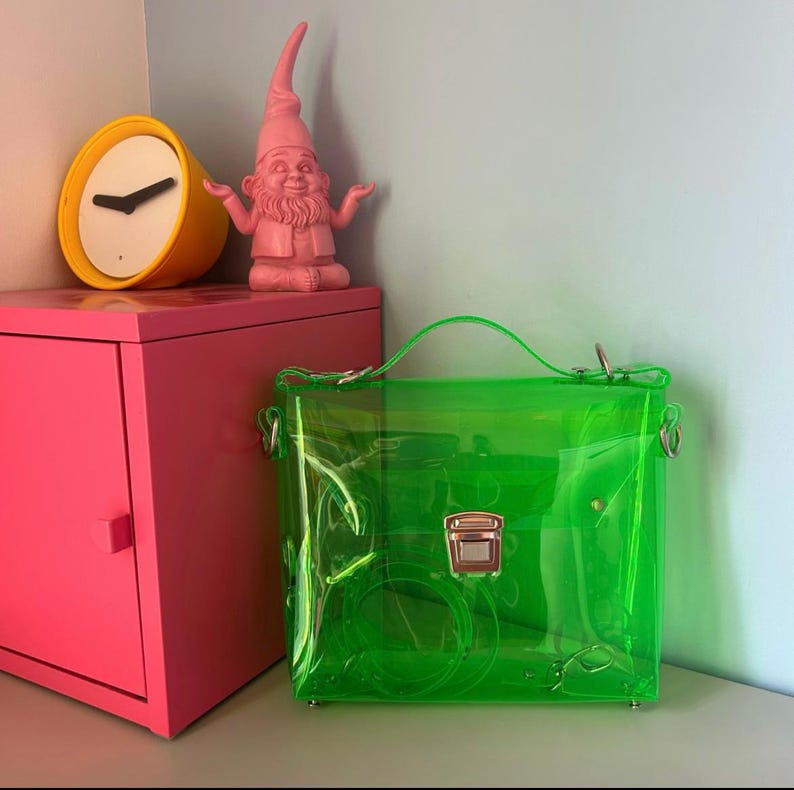 May include: A bright green transparent handbag with a top handle and silver hardware. The bag is next to a pink cabinet with a yellow clock and a pink gnome statue. The background is a light blue wall.