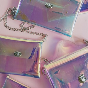 May include: Four iridescent clear plastic purses with silver clasps and a silver chain. The purses are on a pink background.