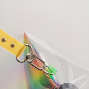 May include: A clear iridescent rainbow bag with a yellow strap and a silver clasp. The bag has a zipper closure and a green plastic loop.