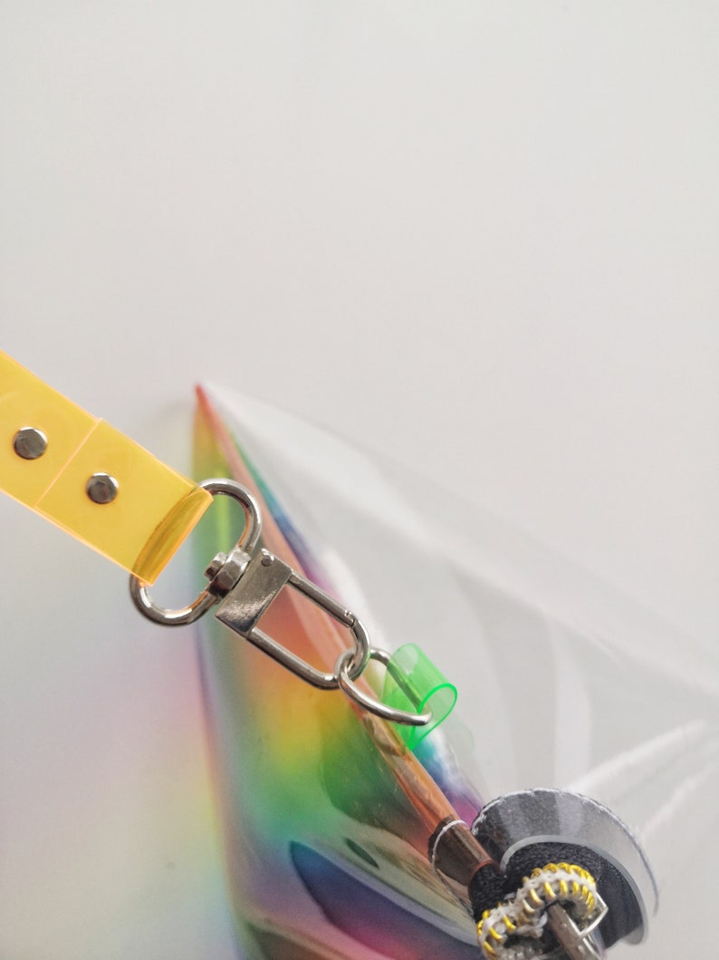 May include: A clear iridescent rainbow bag with a yellow strap and a silver clasp. The bag has a zipper closure and a green plastic loop.