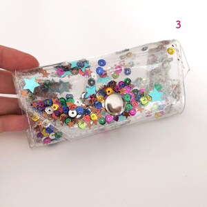 May include: A clear plastic pouch with a silver button closure. The pouch is filled with colorful glitter and sequins, including stars and circles.