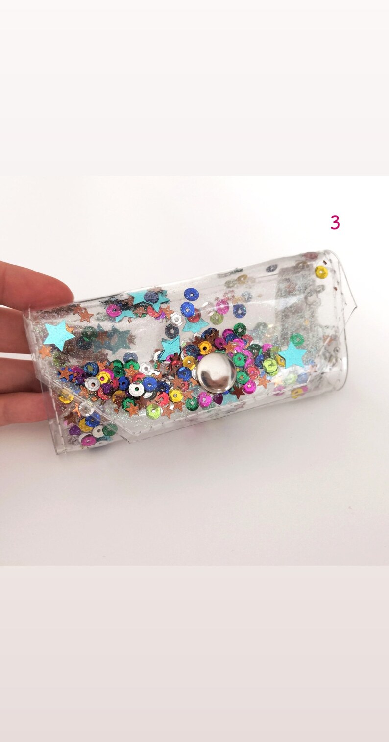 May include: A clear plastic pouch with a silver button closure. The pouch is filled with colorful glitter and sequins, including stars and circles.