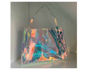 May include: A clear, iridescent tote bag with a rectangular shape and two clear handles. The bag has a shimmering, glittery effect with hues of pink, orange, and blue. The handles are attached to the bag with silver-colored hardware.