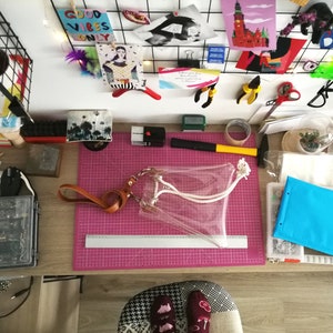 May include: A clear plastic tote bag with a tan leather wrist strap and white rope handles. The bag is positioned on a pink cutting mat next to a silver metal ruler that shows a length of 30 centimeters. The bag features silver metal hardware.