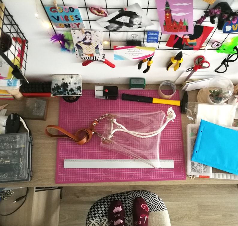May include: A clear plastic tote bag with a tan leather wrist strap and white rope handles. The bag is positioned on a pink cutting mat next to a silver metal ruler that shows a length of 30 centimeters. The bag features silver metal hardware.