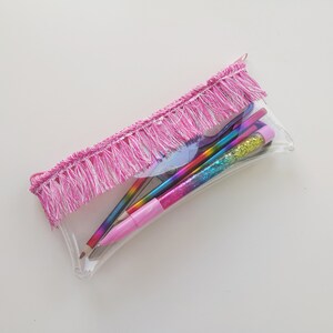 Pu&ograve; includere: Astuccio trasparente con finiture a frange rosa, pieno di matite e penne colorate. Le matite hanno disegni arcobaleno e glitter. L'astuccio &egrave; rettangolare e trasparente, permettendo di vedere il contenuto.