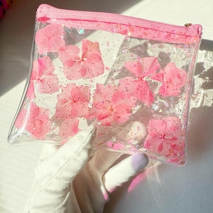 Puede incluir: Una bolsa transparente con cremallera de plástico con flores rosas prensadas y purpurina en el interior.