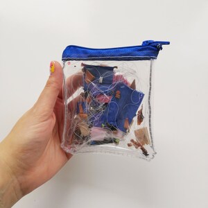 Pode incluir: Uma bolsa transparente e retangular com z&iacute;per e acabamento azul, cheia de retalhos de tecido e linha. A bolsa &eacute; segurada por uma m&atilde;o com um design de unhas colorido. O conte&uacute;do inclui peda&ccedil;os de ganga azul e v&aacute;rios outros restos de tecido.