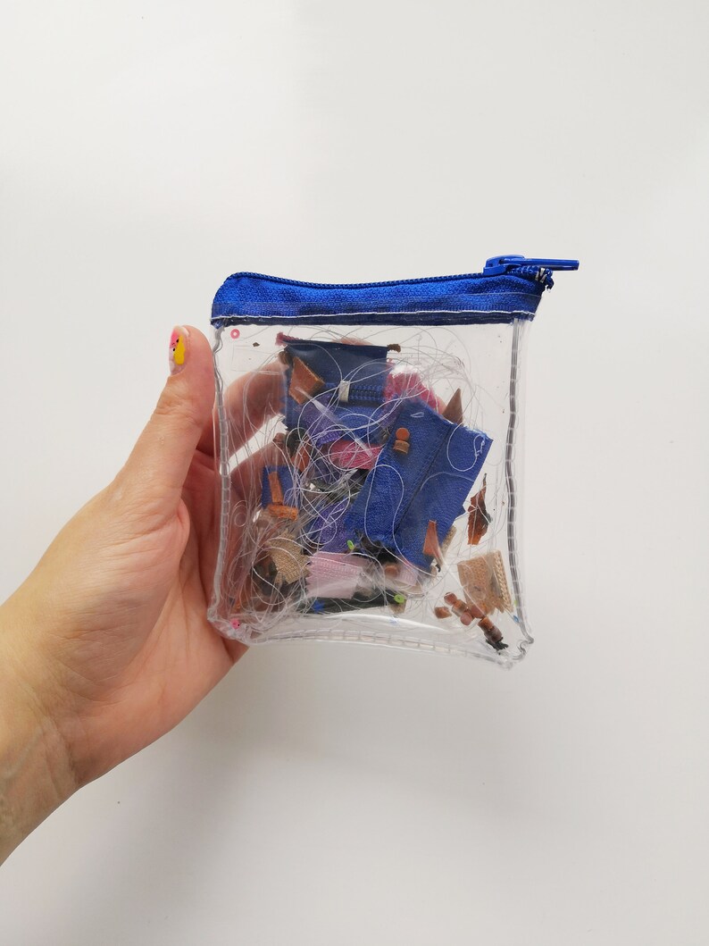 Pode incluir: Uma bolsa transparente e retangular com z&iacute;per e acabamento azul, cheia de retalhos de tecido e linha. A bolsa &eacute; segurada por uma m&atilde;o com um design de unhas colorido. O conte&uacute;do inclui peda&ccedil;os de ganga azul e v&aacute;rios outros restos de tecido.