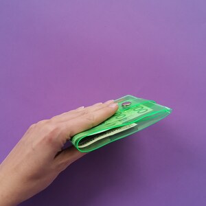 Pu&ograve; includere: Un portafoglio verde neon trasparente con chiusura a bottone automatico argentato, pieno di banconote verdi. Il portafoglio &egrave; tenuto su uno sfondo viola.