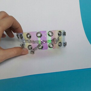May include: A clear choker with a pastel rainbow design. The choker features silver-colored grommets and a small chain with a charm. The accessory is held by a hand against a white and blue background.