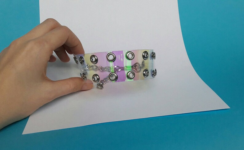 May include: A clear choker with a pastel rainbow design. The choker features silver-colored grommets and a small chain with a charm. The accessory is held by a hand against a white and blue background.