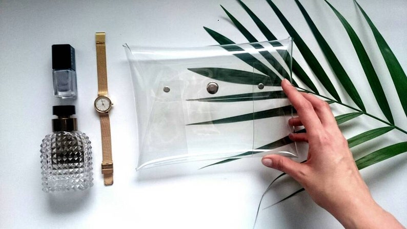 May include: A clear plastic clutch bag with a silver snap closure. The bag is lying on a white surface with a green palm leaf behind it.