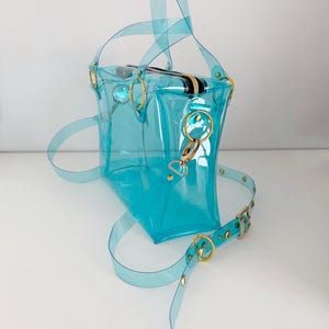 May include: A transparent turquoise handbag with gold-colored hardware. The bag features a top zipper, two handles, and a detachable shoulder strap. The strap is adjustable and has gold-colored metal accents.