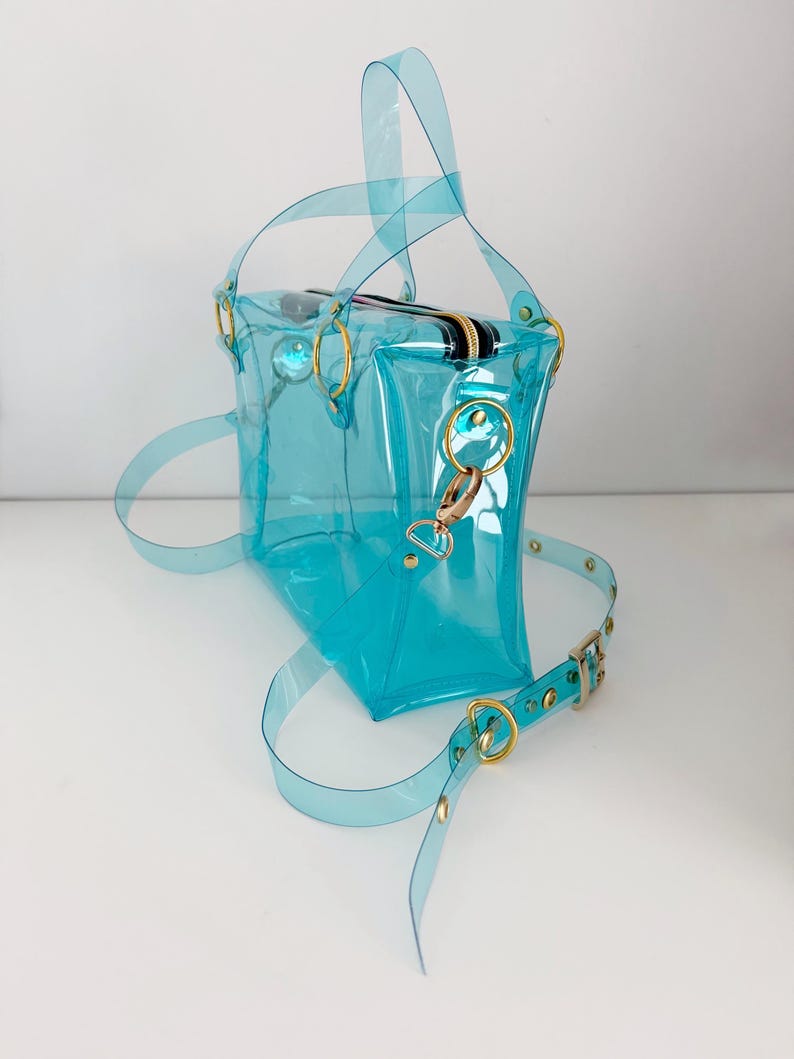 May include: A transparent turquoise handbag with gold-colored hardware. The bag features a top zipper, two handles, and a detachable shoulder strap. The strap is adjustable and has gold-colored metal accents.