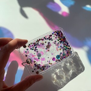 Może przedstawiać: Przezroczysty brelok z plastiku z srebrnym łańcuszkiem i kolorowym brokatem i cekinami w środku.