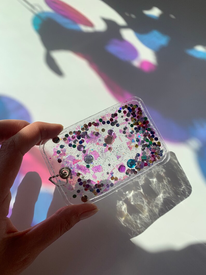 Może przedstawiać: Przezroczysty brelok z plastiku z srebrnym łańcuszkiem i kolorowym brokatem i cekinami w środku.