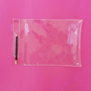 May include: A clear, rectangular pencil case with a black and silver pencil inside, set against a vibrant pink background. The pencil case has a small pocket for the pencil and is secured with silver rivets.