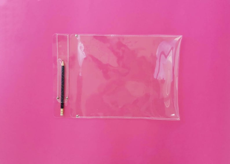 May include: A clear, rectangular pencil case with a black and silver pencil inside, set against a vibrant pink background. The pencil case has a small pocket for the pencil and is secured with silver rivets.