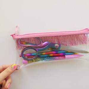 Pu&ograve; includere: Un astuccio trasparente con cerniera e finiture rosa, contenente forbici, matite e penne arcobaleno. Le forbici e le penne hanno una finitura iridescente. L'astuccio &egrave; parzialmente aperto.