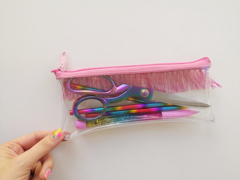 Pu&ograve; includere: Un astuccio trasparente con cerniera e finiture rosa, contenente forbici, matite e penne arcobaleno. Le forbici e le penne hanno una finitura iridescente. L'astuccio &egrave; parzialmente aperto.