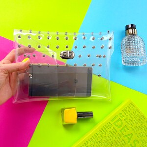 Puede incluir: Un bolso de mano transparente de pl&aacute;stico con tachuelas plateadas y un cierre plateado. El bolso est&aacute; abierto y un tel&eacute;fono inteligente negro est&aacute; dentro. El bolso est&aacute; sobre un fondo rosa, verde y azul.