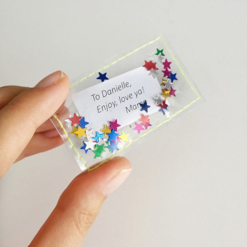 May include: A clear plastic pouch with a yellow stitched border filled with colorful star-shaped confetti. A white card inside the pouch reads "To Danielle, Enjoy, love ya! Mama".